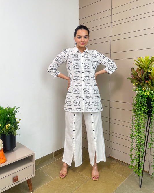 Woman wearing a white outfit with black text standing indoors next to a plant.