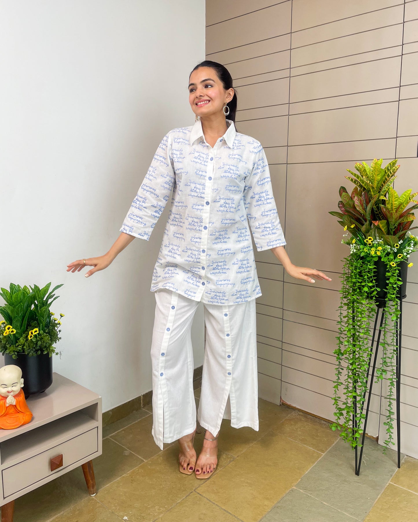 Woman in a white outfit standing in a room with plants and a small table.