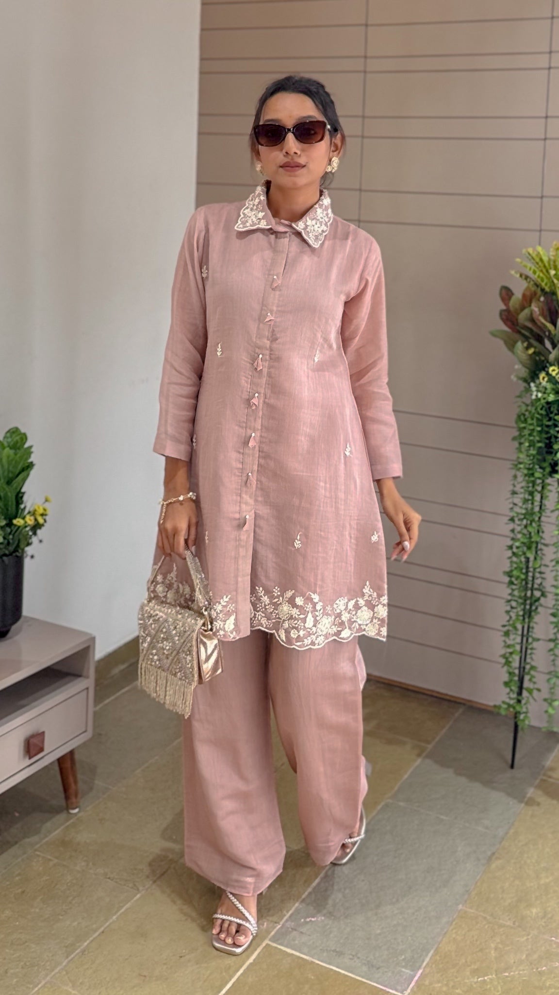 Woman in a dusty rose pink traditional outfit with white embroidery standing indoors.