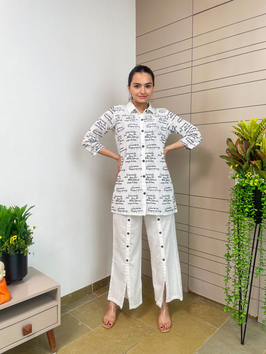 Woman wearing a white outfit with black text standing indoors next to a plant.